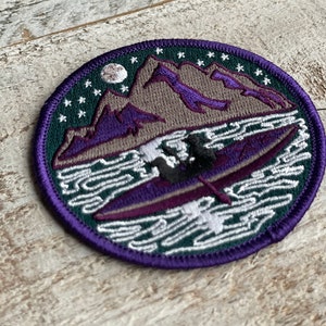 Op de afbeelding: Geborduurde patch met een bergketen, een kajak op het water en een nachthemel met sterren en een maan. De patch heeft een cirkelvorm met een paarse rand en een groene achtergrond.