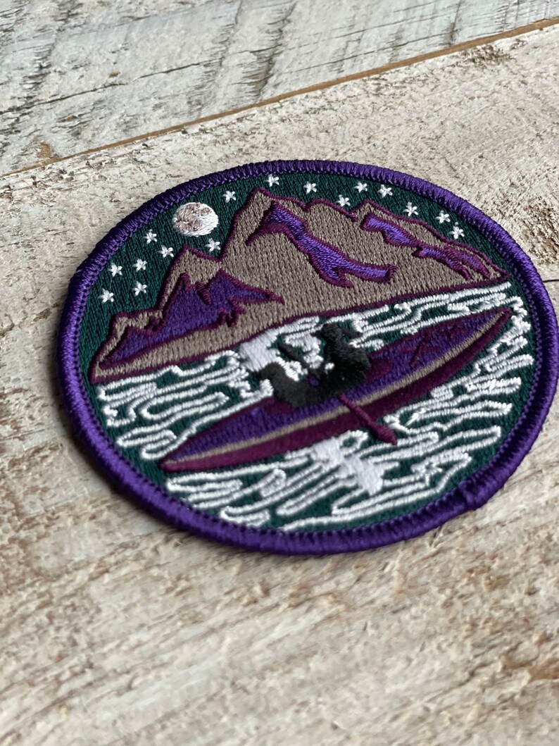 Op de afbeelding: Geborduurde patch met een bergketen, een kajak op het water en een nachthemel met sterren en een maan. De patch heeft een cirkelvorm met een paarse rand en een groene achtergrond.