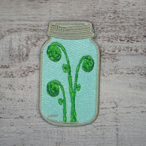 May include: A green and brown embroidered patch of a mason jar with a light blue background and three green fern fronds inside.