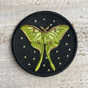 May include: A green luna moth embroidered on a black circular patch with white dots. The patch is perfect for adding a touch of nature to your belongings.