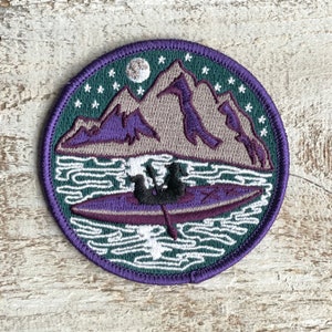 May include: Embroidered patch featuring a mountain scene with a kayak on the water. The design includes purple mountains, a full moon, and stars against a dark green and purple background. The patch is round with a purple border.