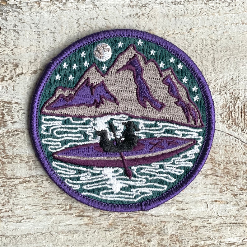 Op de afbeelding: Ronde geborduurde patch met een berglandschap met een kajak. Het ontwerp toont paarse bergen, een volle maan en sterren tegen een donkergroene en paarse achtergrond. De patch heeft een paarse rand.