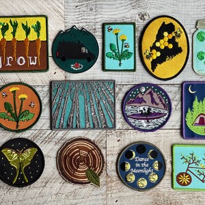 May include: A collection of 12 embroidered iron-on patches featuring nature-inspired designs. The patches include a dandelion, a forest, a mountain range, a moon, a bicycle, a jar of ferns, a moth, a tree stump, a beehive, a tent, and a row of carrots with the word "grow" written below.