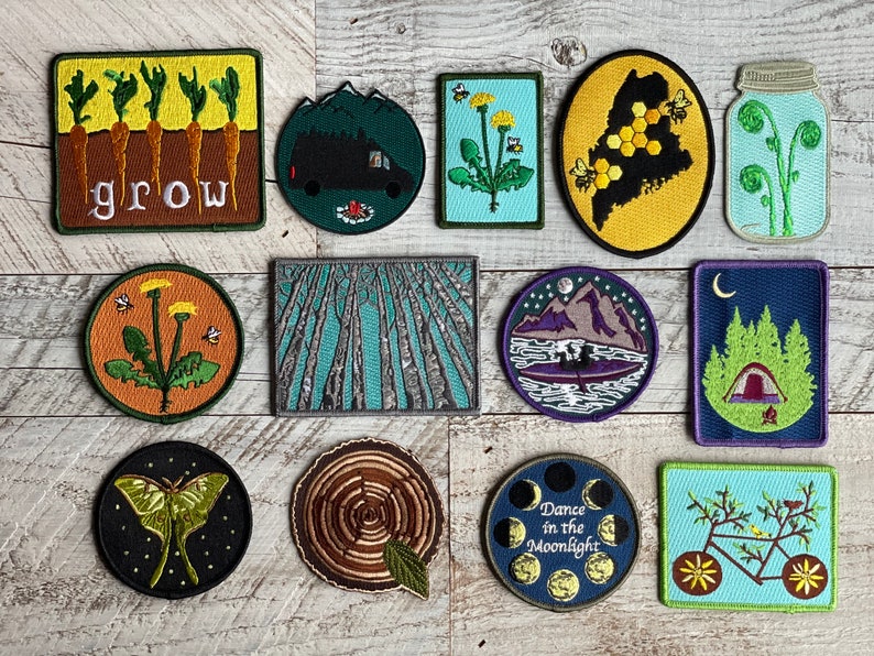 May include: A collection of 12 embroidered iron-on patches featuring nature-inspired designs. The patches include a dandelion, a forest, a mountain range, a moon, a bicycle, a jar of ferns, a moth, a tree stump, a beehive, a tent, and a row of carrots with the word "grow" written below.