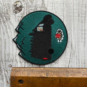 May include: A black and green embroidered patch with a black silhouette of a creature with a red beak and a white and red flower design. The patch is circular and has a jagged edge.