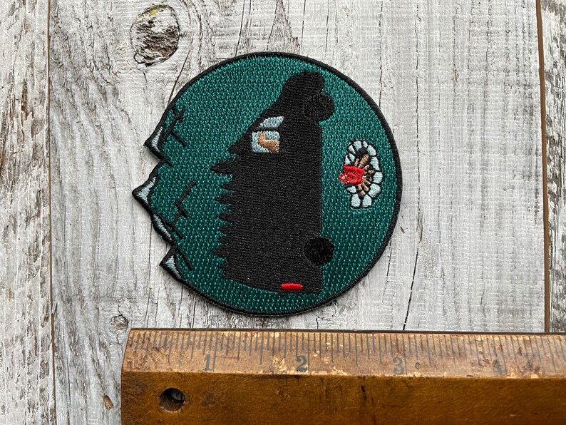 May include: A black and green embroidered patch with a black silhouette of a creature with a red beak and a white and red flower design. The patch is circular and has a jagged edge.