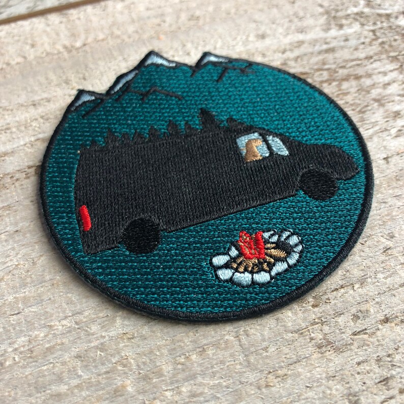 May include: A teal blue embroidered patch with a black van in front of a mountain range. The van has a window with a person inside. There is a campfire in front of the van.