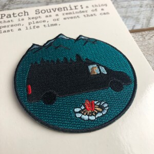 May include: A teal blue embroidered patch with a black van driving through a forest with a campfire in the foreground. The text "Patch Souvenir: a thing that is kept as a reminder of a person, place, or event that can last a life time." is printed on the background.