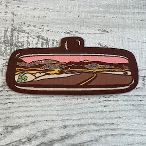 May include: A brown and beige embroidered patch depicting a desert landscape with a pink sunset, a winding road, and a small green bush in the foreground.