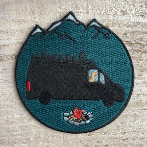 May include: Embroidered patch featuring a black van with a campfire in front of it. The van is silhouetted against a teal blue background with white mountains in the distance.