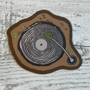 May include: A brown fabric patch with a stylized image of a record player. The record is a tree trunk with rings, and the needle is a branch with a green leaf. There are small birds flying around the record.