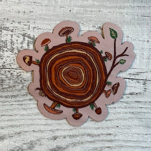 May include: A brown and beige fabric patch with a cross-section of a tree trunk and mushrooms around it. The tree trunk has a brown and beige swirl pattern. The mushrooms are brown and beige with white spots.
