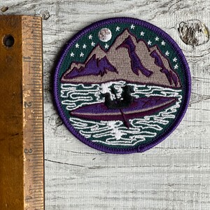 Op de afbeelding: Een ronde geborduurde patch met een berglandschap met een meer en een kano. De patch heeft een donkergroene rand met witte sterren en een maan. De bergen zijn paars en grijs, en de kano is paars.