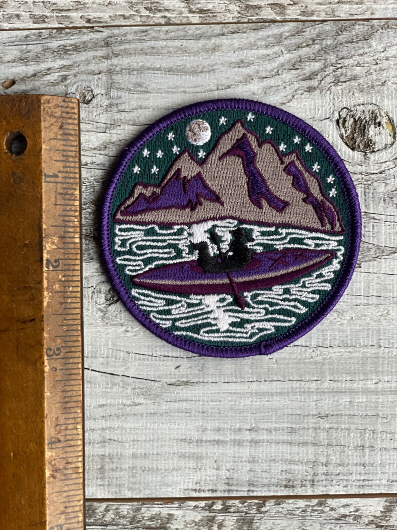 Op de afbeelding: Een ronde geborduurde patch met een berglandschap met een meer en een kano. De patch heeft een donkergroene rand met witte sterren en een maan. De bergen zijn paars en grijs, en de kano is paars.