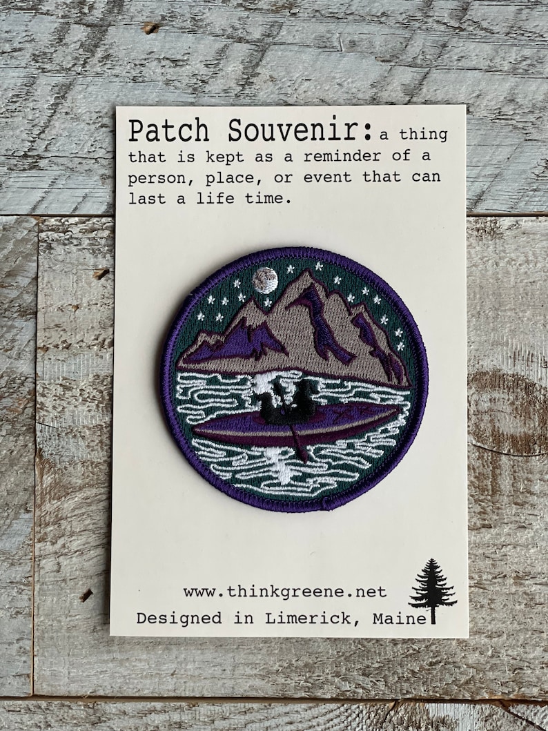Op de afbeelding: Ronde souvenirpatch met een berg- en meerontwerp in paars, grijs en groen. De patch heeft een witte rand en bevat de tekst "Patch Souvenir" en "Designed in Limerick, Maine."