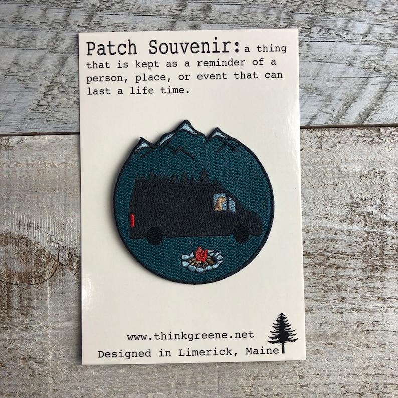 May include: A round, green embroidered patch with a black van silhouette, a campfire, and mountains in the background. The text "Patch Souvenir: a thing that is kept as a reminder of a person, place, or event that can last a life time." is printed on a white card behind the patch. The text "www.thinkgreene.net Designed in Limerick, Maine" is printed below the patch.