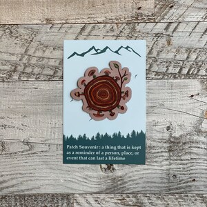 May include: A brown and beige patch with a tree stump design. The stump has rings and is surrounded by mushrooms and leaves. The patch has a white background with a blue mountain range at the top. The text "Patch Souvenir: a thing that is kept as a reminder of a person, place, or event that can last a lifetime" is printed below the patch.