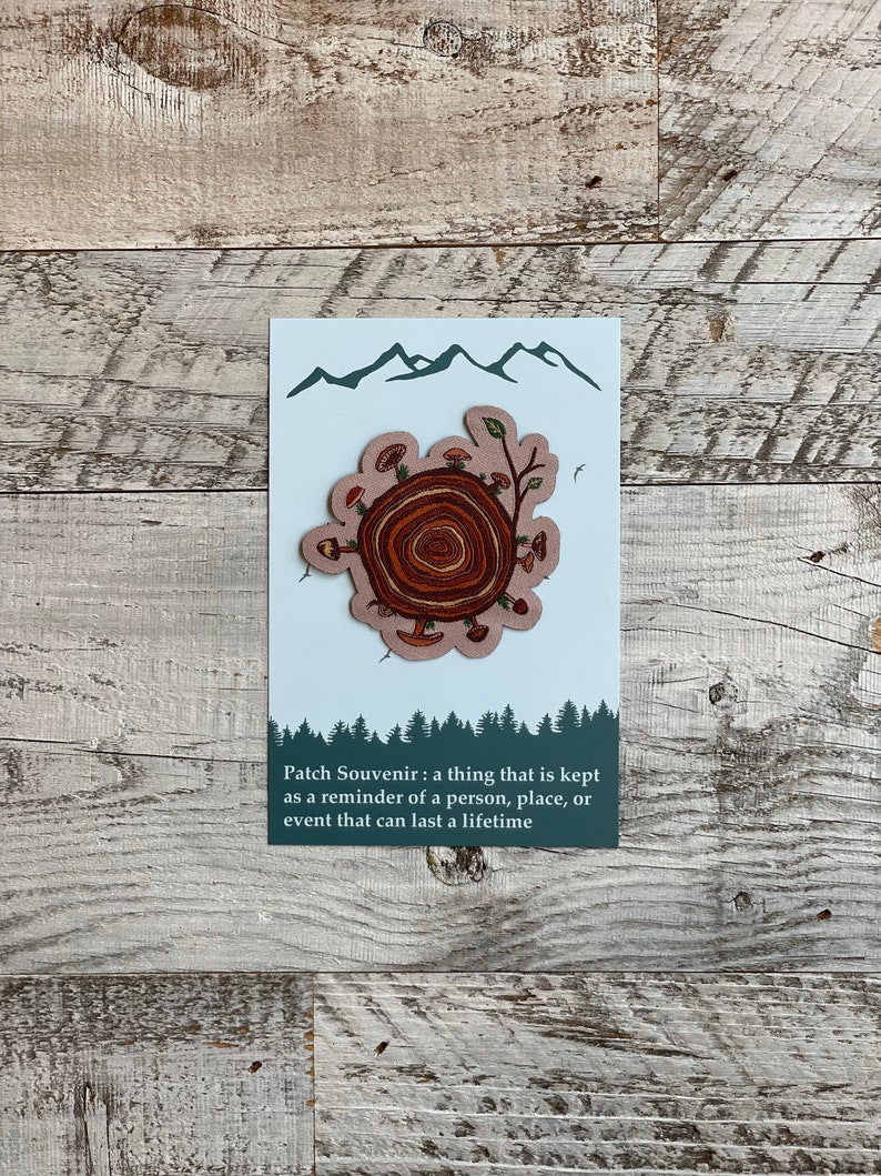 May include: A brown and beige patch with a tree stump design. The stump has rings and is surrounded by mushrooms and leaves. The patch has a white background with a blue mountain range at the top. The text "Patch Souvenir: a thing that is kept as a reminder of a person, place, or event that can last a lifetime" is printed below the patch.
