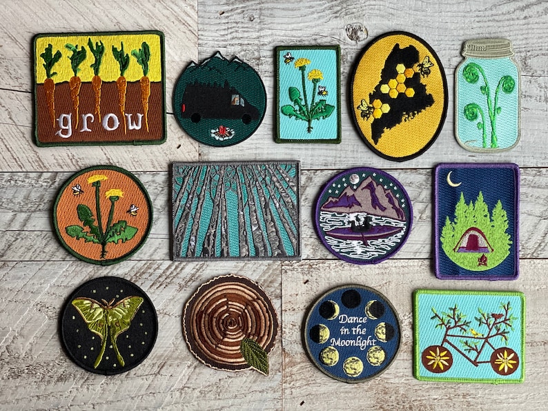 Op de afbeelding: Een verzameling geborduurde patches met ontwerpen met een natuurthema. De patches tonen afbeeldingen van wortels met het woord "grow", een camper, paardenbloemen, een kaart van Maine met bijen, varens, bomen, bergen, een tent, een mot, een boomdoorsnede, een maanfasedesign en een fiets.