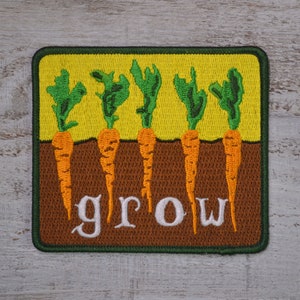 May include: A green and brown embroidered patch with five orange carrots growing out of the ground. The word "grow" is stitched in white letters below the carrots.