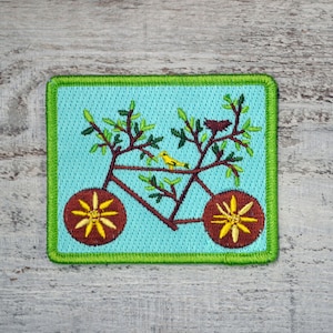 May include: Embroidered patch featuring a bicycle design with tree branches and a yellow bird. The bicycle wheels are brown with yellow floral accents. The background is light blue, framed by a green border.