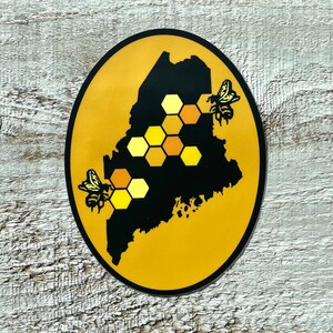 May include: A yellow circle sticker with a black outline featuring a stylized map of Maine with a honeycomb pattern and two bees.