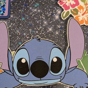 May include: A blue cartoon character with large eyes and a wide smile. The character is peeking out from behind a glittery blue background with white and gold glitter. The character has a black nose and a light blue body.