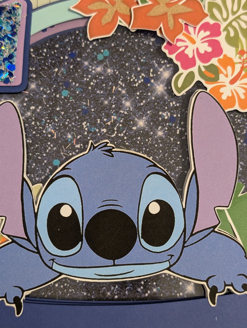 May include: A blue cartoon character with large eyes and a wide smile. The character is peeking out from behind a glittery blue background with white and gold glitter. The character has a black nose and a light blue body.