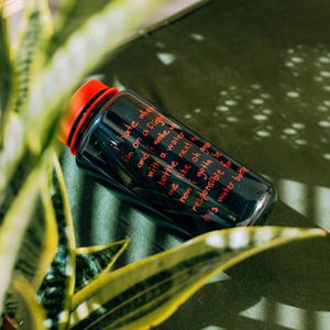 May include: A dark blue water bottle with a red cap, resting on a green surface. The bottle has red text that reads, "I hate when I'm on a flight and I wake up with a water bottle next to me..." The bottle is surrounded by green plants.