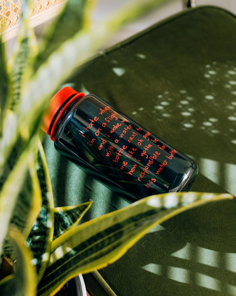 May include: A dark blue water bottle with a red cap, resting on a green surface. The bottle has red text that reads, "I hate when I'm on a flight and I wake up with a water bottle next to me..." The bottle is surrounded by green plants.