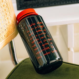 May include: Black water bottle with a red lid. The bottle has red text that reads "I hate when I'm on a flight and I wake up with a water bottle next to me..." The bottle is on a green surface.