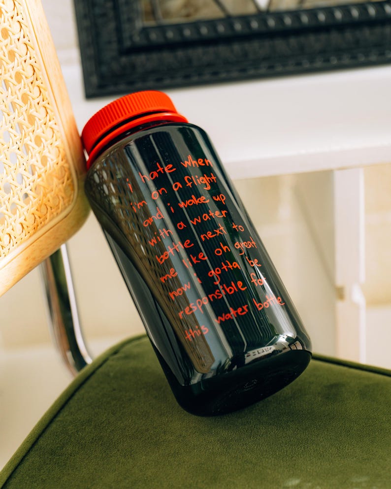 May include: Black water bottle with a red lid. The bottle has red text that reads "I hate when I'm on a flight and I wake up with a water bottle next to me..." The bottle is on a green surface.