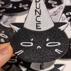 Op de afbeelding: Zwart-witte geborduurde strijkpatch van een kat met een narrenmuts. De kat heeft een chagrijnige uitdrukking en de narrenmuts zegt "DUNCE" in zwarte letters.