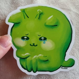 May include: A green sticker depicting a cartoon creature with a sorrowful expression. The creature has large, tearful eyes, small antennae, and a small mouth. The sticker has a white border and the name "Mussu" is printed on the bottom right.