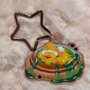以下が含まれることがあります： 緑色の浴槽の中に黄色いアヒルの子が描かれた木製のキーホルダー。キーホルダーは銀色の星型のリングに繋がっています。