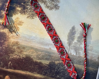 May include: A handmade friendship bracelet with a red, green, and white geometric pattern. The bracelet has a braided extension with a tassel of colorful threads. The bracelet is displayed on a vintage painting.