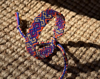 May include: A handmade friendship bracelet with a woven pattern in shades of blue, orange, and yellow. The bracelet is knotted at the ends with a tassel. The bracelet is resting on a textured, brown surface.