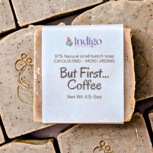 May include: A light brown, speckled exfoliating soap bar wrapped in a white paper label. The label features the text 'Indigo SOAPWORKS', '97% Natural small batch soap', 'EXFOLIATING - MOISTURIZING', 'But First... Coffee', and 'Net Wt 4.5-5oz'.