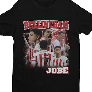 Può includere: Maglietta nera con una grafica in stile vintage di giocatori di calcio. La grafica presenta il testo "Bellingham" e "Jobe" in uno schema di colori rosso e bianco.