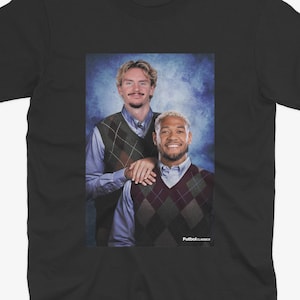 May include: Black t-shirt featuring a portrait of two men wearing argyle vests over button-down shirts. The image has a blue background and the text "FutbolClassics" is visible.