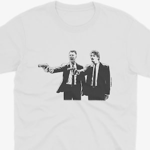 Puede incluir: Camiseta blanca con un gráfico en blanco y negro de dos hombres con trajes apuntando con pistolas. La ilustración es una representación estilizada de una escena de una película. La camiseta está hecha de un material suave y cómodo.