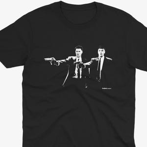 Pode incluir: Camiseta preta com um gráfico branco que retrata dois homens de terno segurando armas. A ilustração é um design estilizado e monocromático. A camiseta tem gola redonda e um estilo clássico.