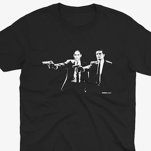 Puede incluir: Camiseta negra con un gráfico blanco de dos hombres con traje apuntándose con pistolas. El texto "Pulp Fiction" está impreso debajo del gráfico.