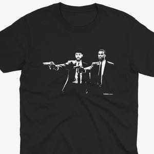 May include: Black t-shirt featuring a black and white graphic of two men in suits holding guns. The image is a stylized illustration, with the figures in a dynamic pose. The shirt is a classic crew neck style.