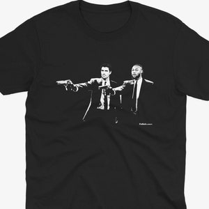 Può includere: T-shirt nero e bianco con una grafica che rappresenta due uomini in giacca e cravatta che tengono delle pistole, con il testo "Pulp Fiction" in basso.