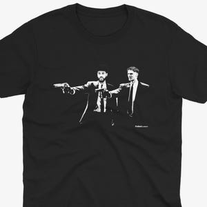 May include: Black t-shirt featuring a black and white graphic of two men in suits pointing guns. The image is a stylized illustration, with the figures in a classic pose. The shirt is a crew neck style.