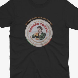 Pu&ograve; includere: Maglietta nera con una grafica circolare di un uomo che tiene una birra. La grafica dice "Sandro Tonali" e "Centrocampista italiano premium".