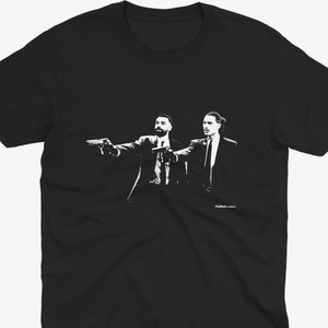May include: Black t-shirt featuring a white graphic of two men in suits pointing guns. The image is a stylized illustration. The text "Folkdances" is printed in white at the bottom right of the shirt.