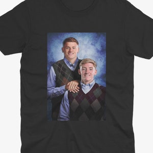 May include: A black t-shirt featuring a photograph of two men wearing sweaters. The men are standing side-by-side and smiling. The background is a blue and white gradient.
