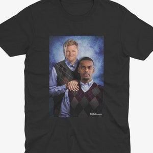 May include: A black t-shirt with a photo of two men wearing sweaters. The man on the left is wearing a blue shirt and a brown and white patterned sweater. The man on the right is wearing a blue shirt and a red and white patterned sweater. The background is a blue and white gradient.